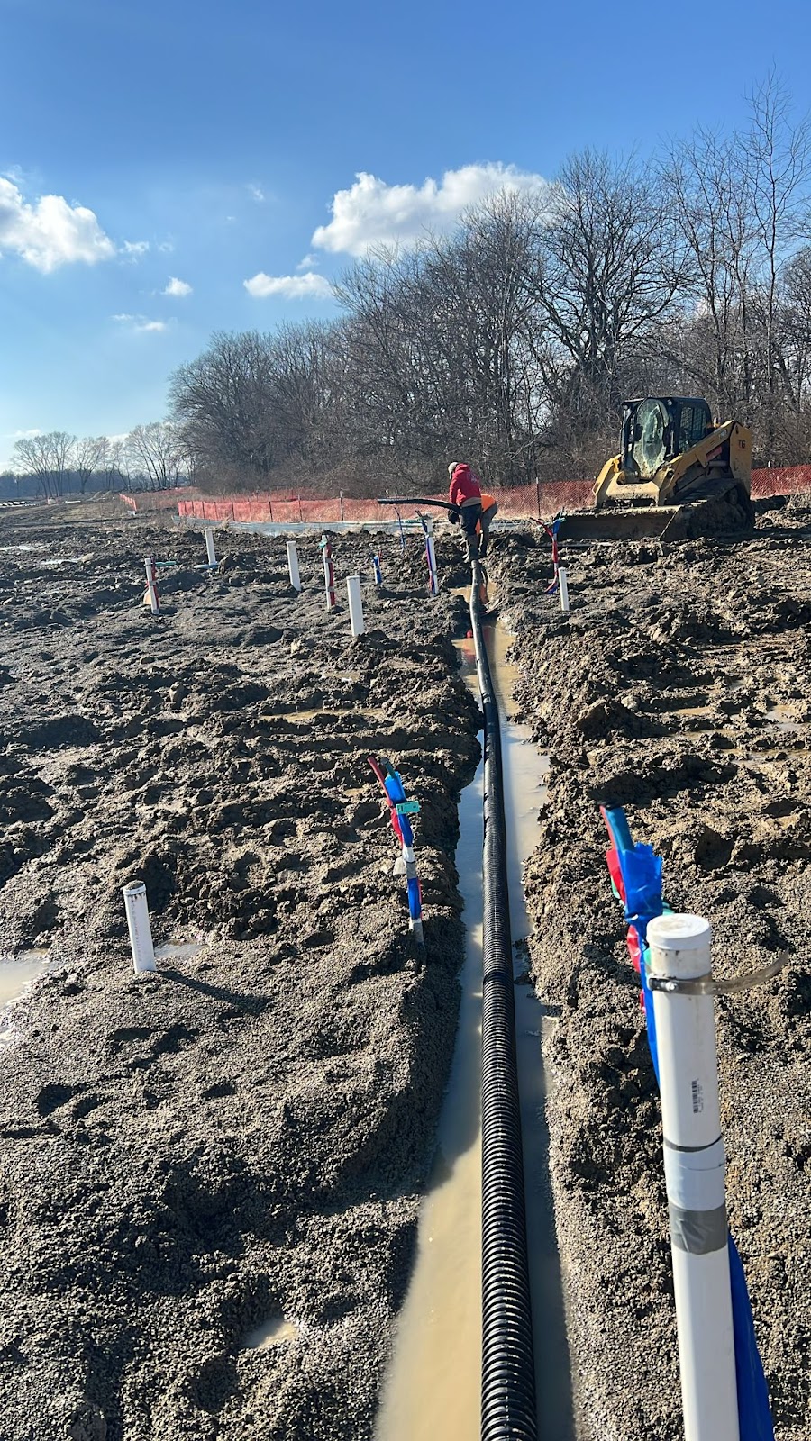 A Mendez Plumbing crew member laying drain line in a trench on a new build site under a blue sky